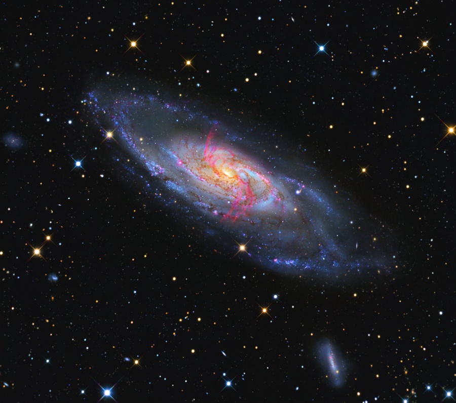 Eine Spiralgalaxie mit blauen Spiralarmen ist von Sternen und Galaxien umgeben. Vor der Galaxie verlaufen sehr auffällige rote Filamente.