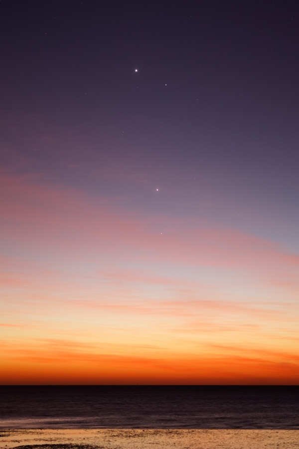 Am Himmel leuchten in der Dämmerung über einem Gewässer drei Himmelslichter. Der Himmel verläuft von orangefarben über hellgelb bis dunkelviolett, er ist von zarten rötlichen Wolken überzogen.