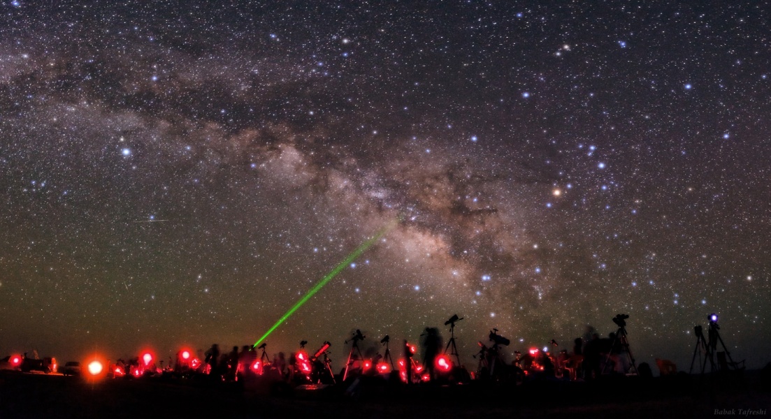Unter einem sternklaren Himmel mit Milchstraße sind Silhouetten von Sternfreunden und rote Lämpchen zu sehen, aus der Menge zielt ein grüner Lichtstrahl schräg nach oben.