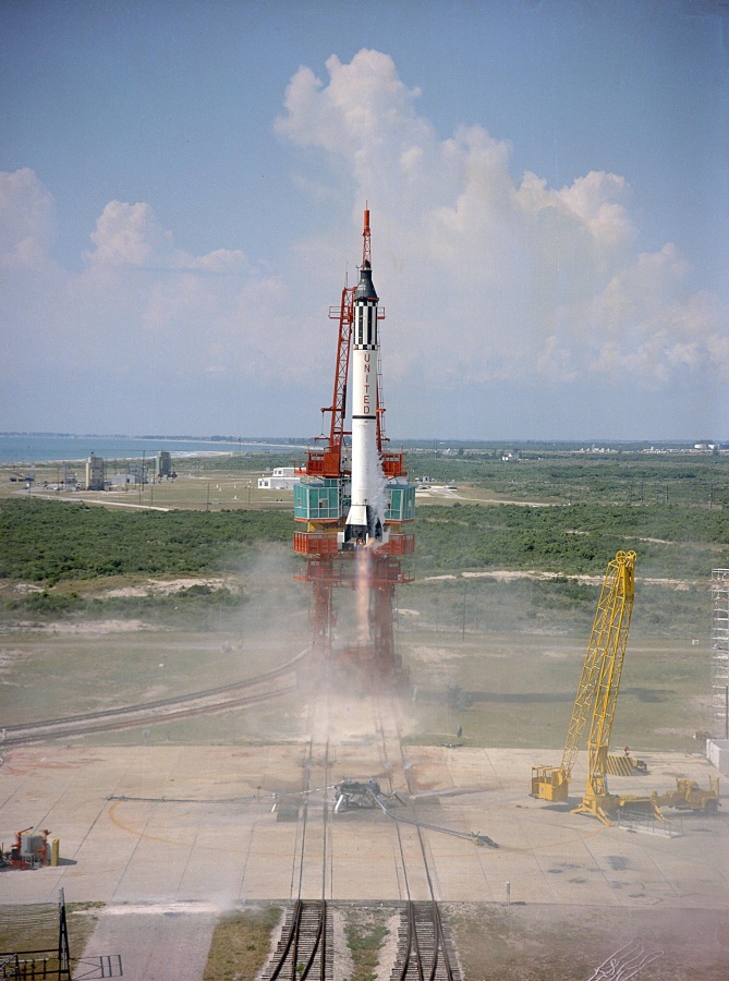 Mitten im Bild startet eine Rakete, am Boden sind Schienen zu sehen, rechts steht ein gelber Kran, im Hintergrund verläuft eine Küstenlinie vor dem Meer.