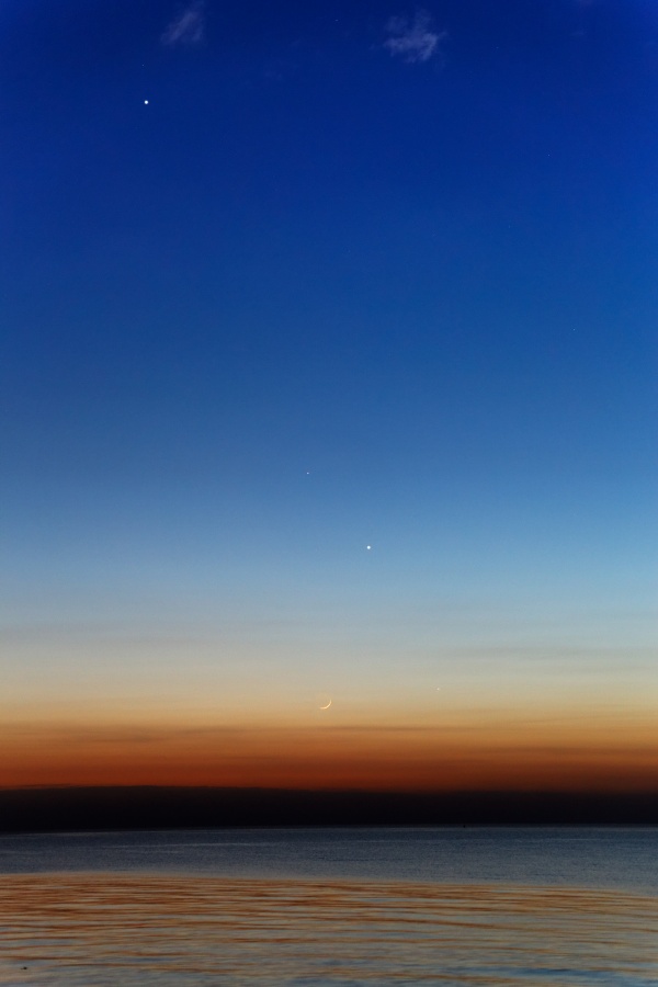 Über einem Gewässer, das am Horizont von einem dunklen Streifen begrenzt ist, über dem der Himmel mit braunrot beginnt, dann in hellgelb übergeht und nach oben hin dunkelblau verläuft, schwegen ein Sichelmond und helle Planeten.