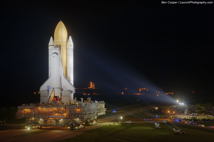 In der Nacht wird die Raumfähre Atlantis zur Startrampe ausgerollt, sie befindet sich links auf dem Raupenfahrzeug, von rechts leuchten Scheinwerfer zur Raumfähre.