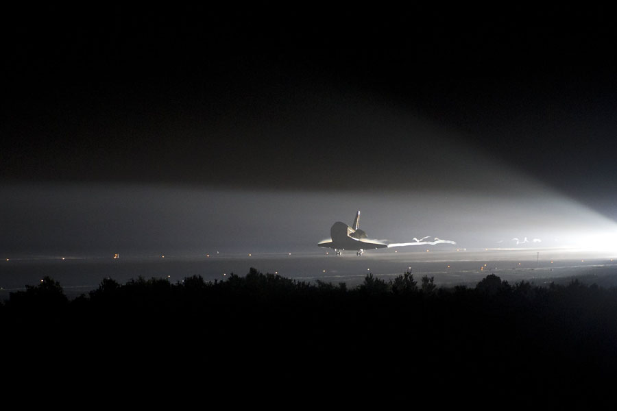 Die Raumfähre Endeavour landet nachts, von links leuchten Scheinwerfer auf den Spaceshuttle, der Rest des Bildes ist dunkel.