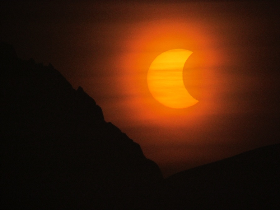 Im Bild leuchtet eine orangefarbene sichelförmige Sonne hinter zarten Wolkenstreifen.