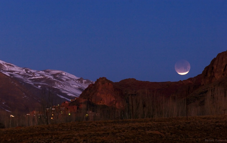 Über einem Gebirge leuchtet eine Mondsichel, es ist der Vollmond, der teilweise vom Erdschatten verdunkelt ist. Der untere Teil des Mondes ist hell.