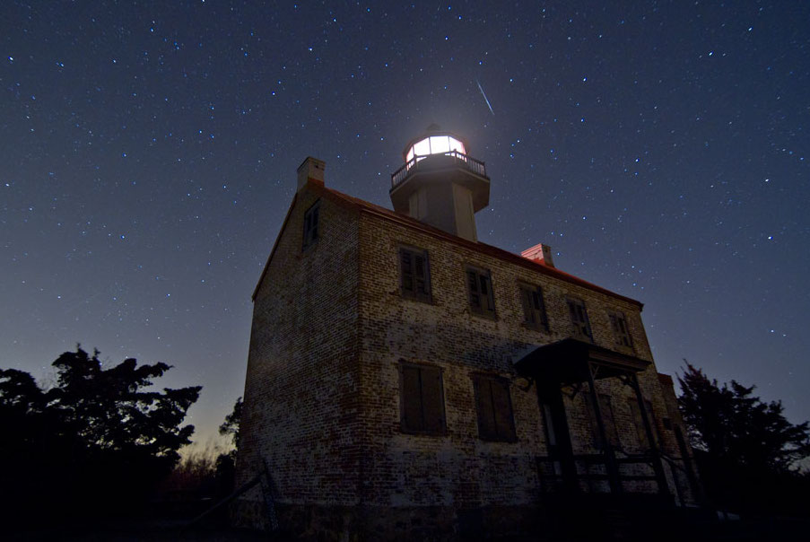 Auf einem düster wirkenden Gebäude steht ein Durm mit einem beleuchteten Glasaufbau. Das Gebäude ist von Bäumen umgeben. Das Bild wurde in der Nacht fotografiert, am dunklen Himmel sind Sterne und ein Meteor zu sehen.