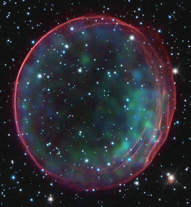 Der Nebel füllt fast das ganze Bild. Außen ist ein roter Ring, der an eine Seifenblase erinnert, innen sind einige grün schimmernde Nebelflecke. Das Bild ist voller weißer Sterne.