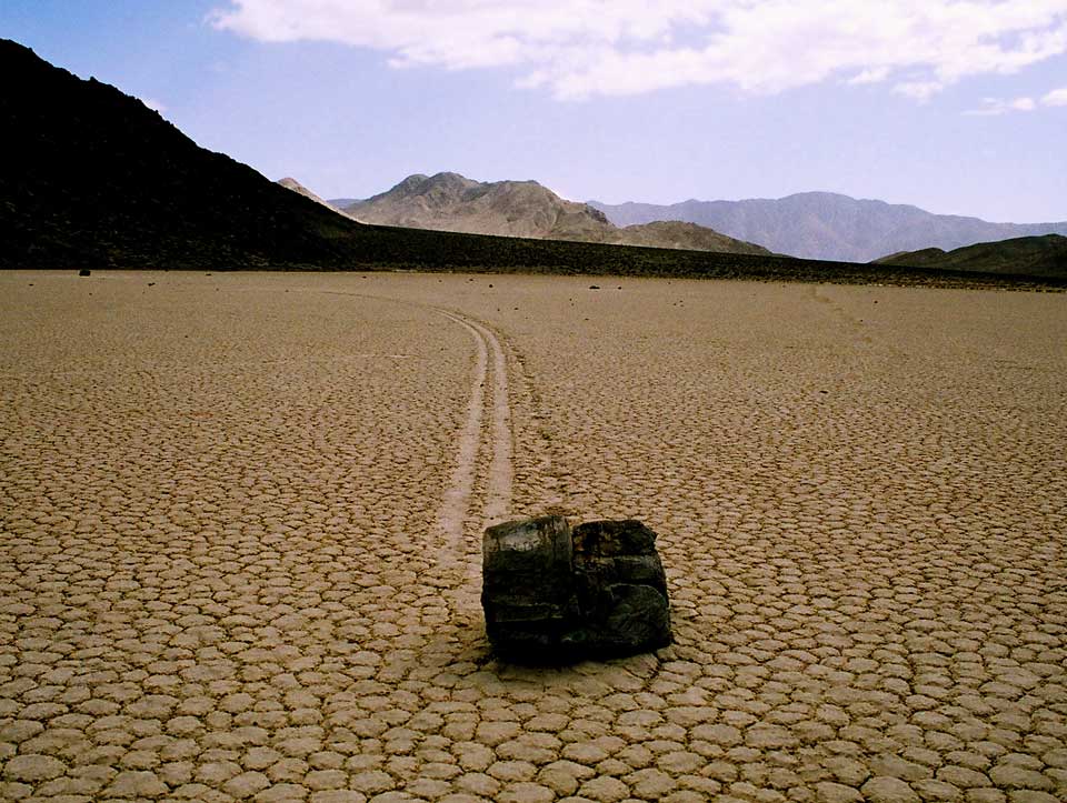 Vor einer gebirgigen Landschaft ist eine große Ebene aus rissiger Erde. Ein dunkler Stein, der vorne liegt, hat eine breite Spur über die rissige Ebene gezogen.