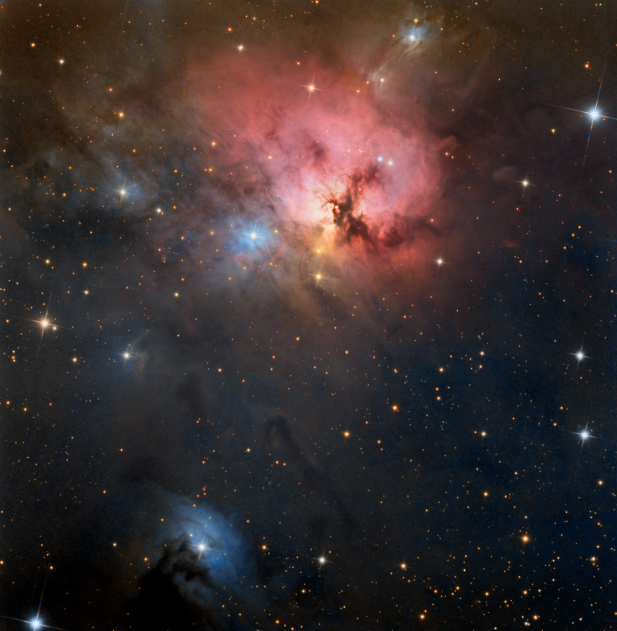 Oben leuchtet ein roter Nebel mit dunklen Ästen davor, der entfernt an den Trifidnebel erinnert. Im Bild sind mehrere Sterne mit blau leuchtenden Nebeln verteilt.