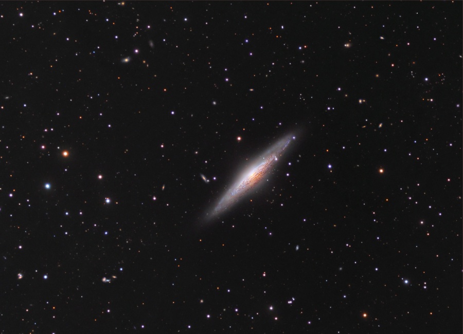 Schräg im Bild liegt eine Galaxie, die fast von der Seite sichtbar ist. Ihre Staubbahnen sind markant vor den hellen Sternen erkennbar. Im Hintergrund sind sterne und Galaxien verteilt.