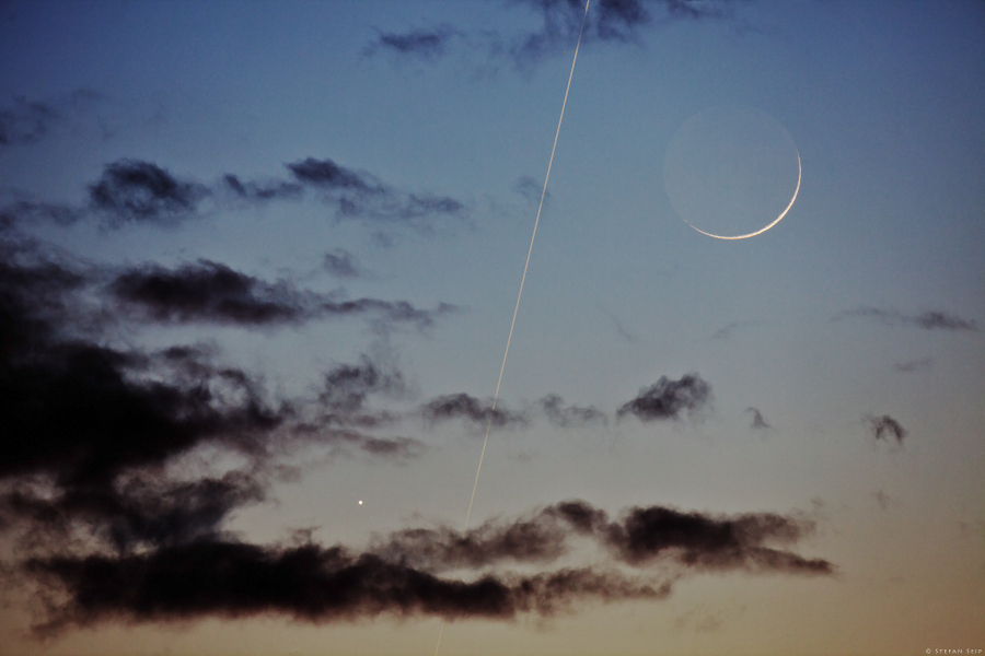 Hinter dunklen Wolken leuchten in der Dämmerung links unten der Planet Merkur, rechts oben der zarte Sichelmond, dazwischen zieht die ISS eine Leuchtspur.