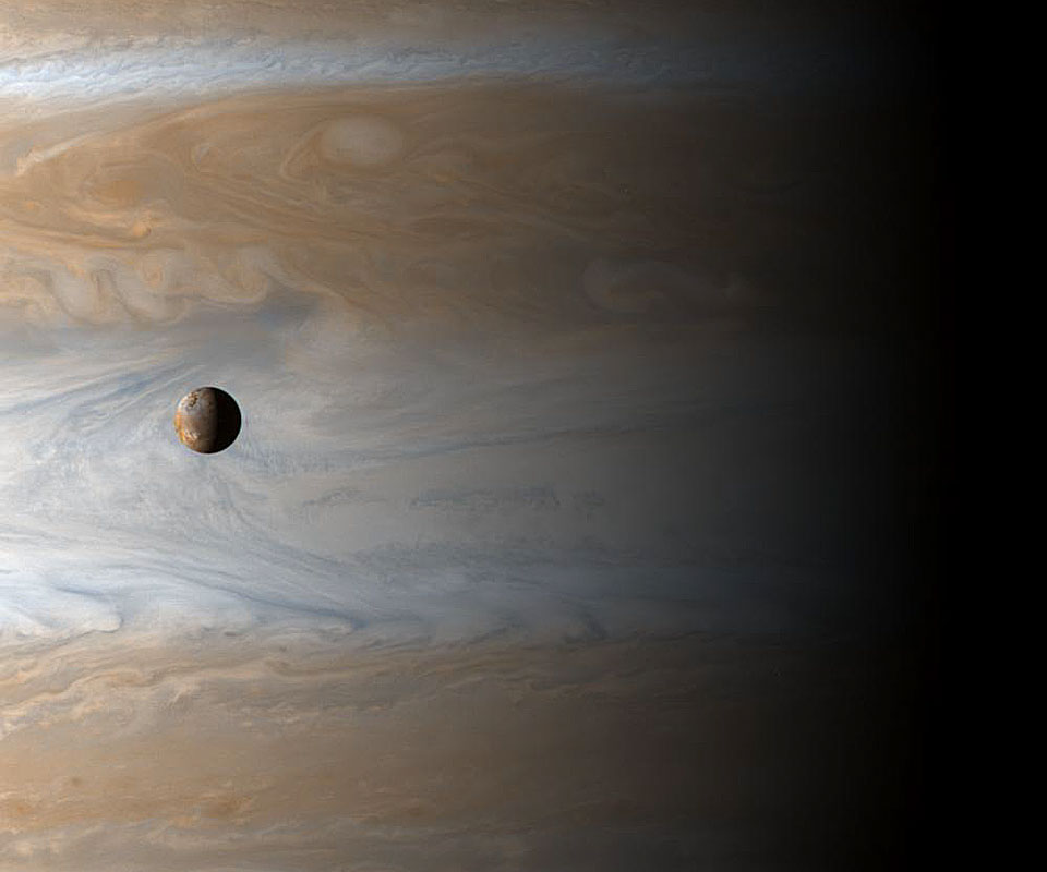 Vor den Wolkenoberflächen von Jupiter schwebt der kleine kugelige Mond Io. Auf beiden Himmelskörpern ist die Licht-Schattengrenze erkennbar, auf dem riesigen Jupiter verläuft sie sehr diffus.
