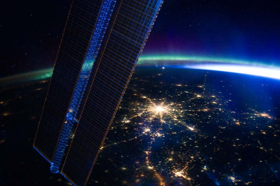 Hinter einem Solarpaneel sieht man die Erde, in der Mitte leuchtet die russische Hauptstadt Moskau wie ein heller Stern. Am Horizont ist ein türkis leuchtendes Polarlicht erkennbar.