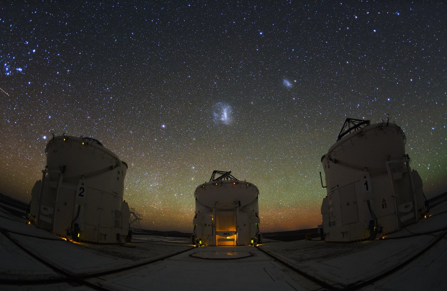 In der Abenddämmerung stehen drei Hilfsteleskope, darüber leuchten die Magellanschen Wolken am sternklaren Himmel. Von links ragt das Sternbild Orion ins Bild.