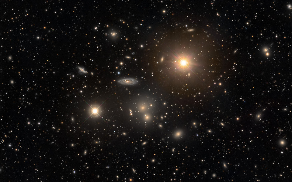 Im Bild leuchten zwei helle Stern und mehrere helle Galaxien, dahinter sind viele kleine Galaxien wie Sterne verteilt. 