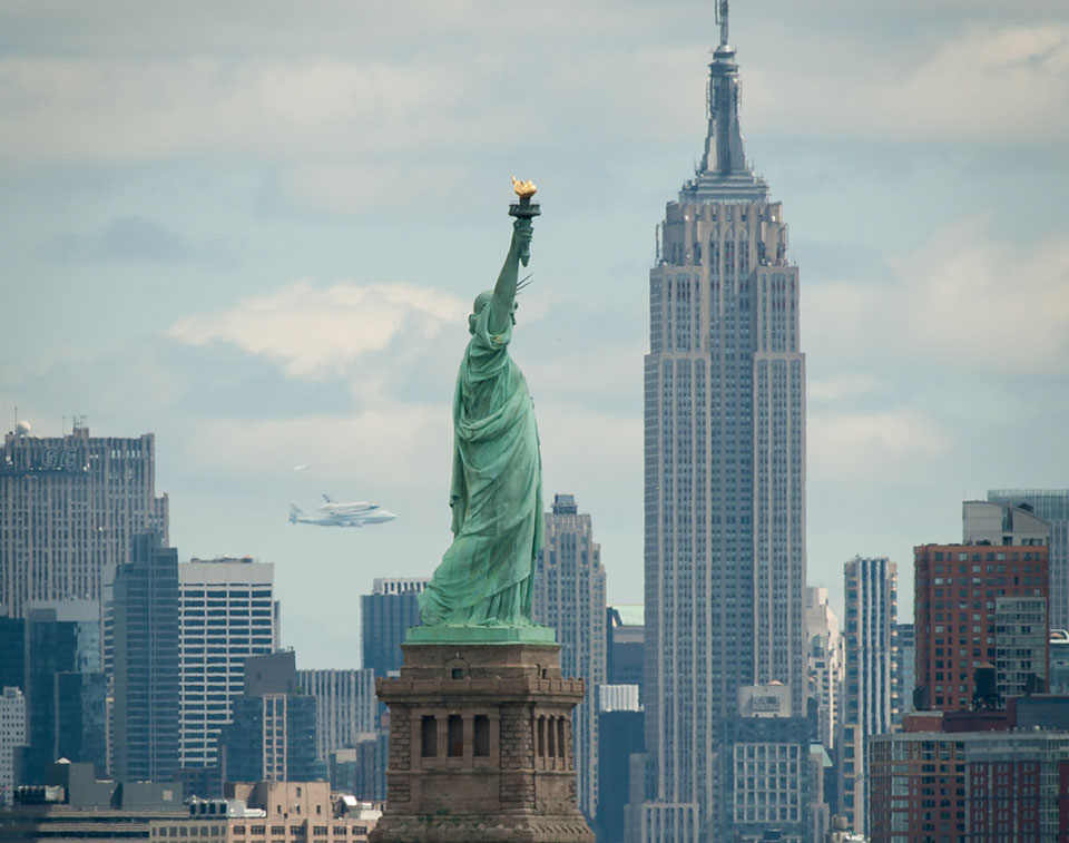 Die Skyline von New York zeigt in der Mitte die Freiheitsstatue und das Empire State Building. Links neben den beiden sieht man am Horizont eine Boeing 747 mit der Test-Raumfähre Enterprise.