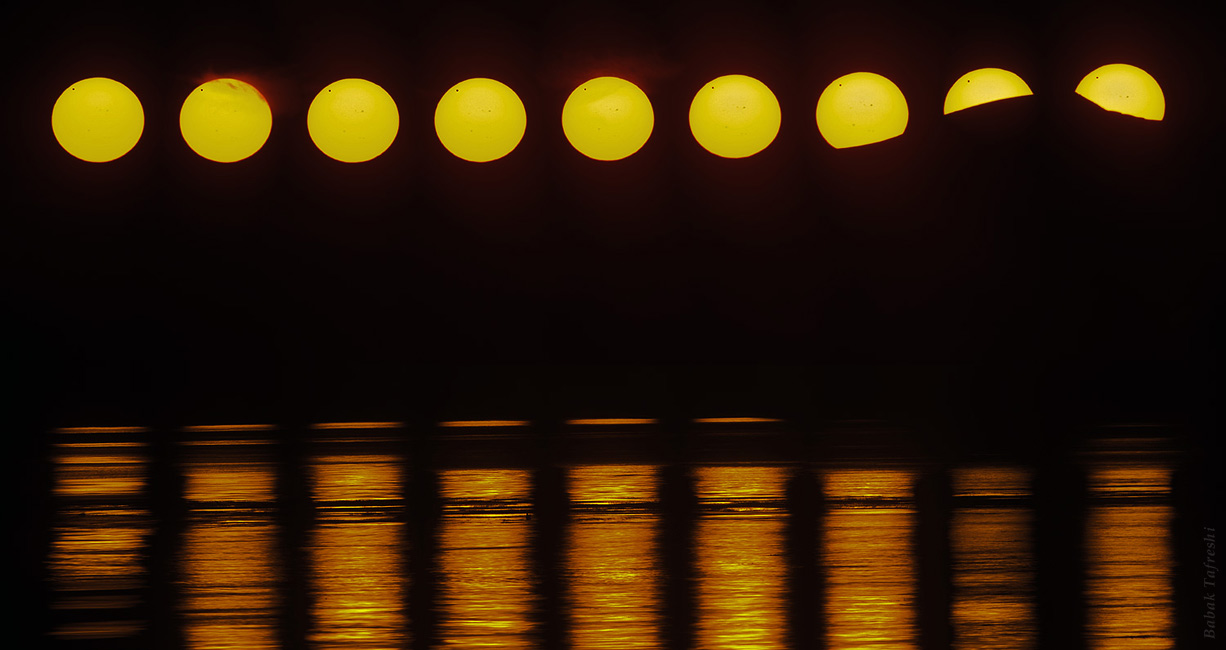 Über einem Gebirge zieht die Sonne waagrecht den Horizont entlang und verschwindet rechts teilweise hinter einem Berg. Oben zieht ein dunkler Punkt über die Sonnenscheibe, es ist der Planet Venus. Die Sonne spiegelt sich vorne im Meer.
