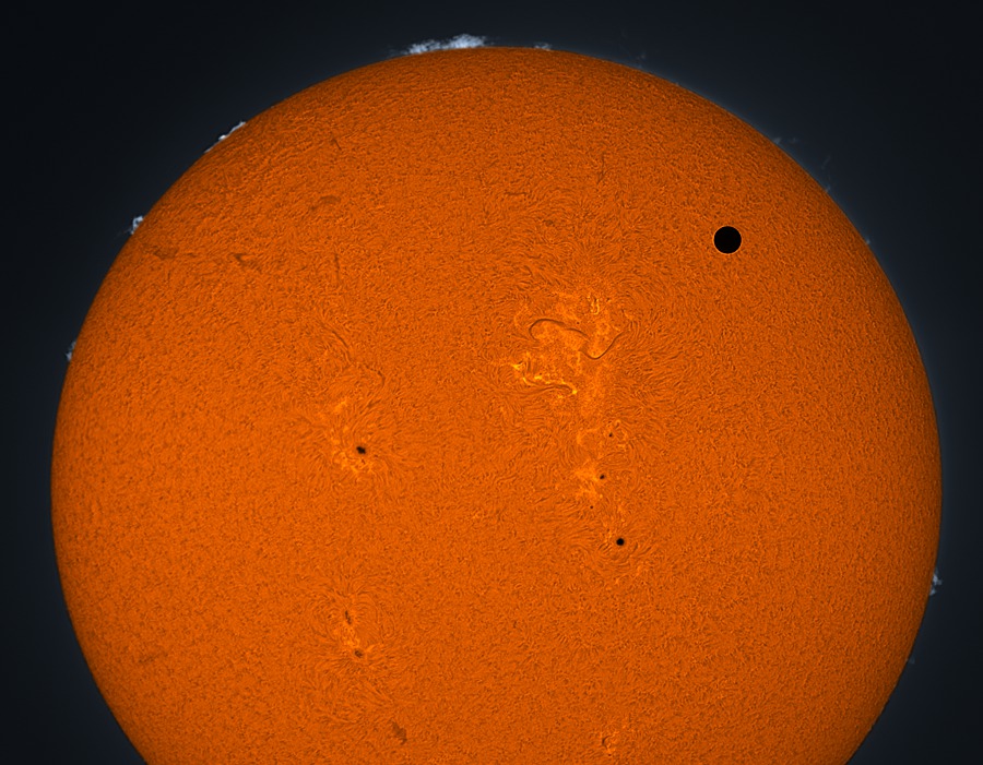 Vor der orangeroten Sonne mit Granulation, Sonnenflecken und Filamenten ist rechts oben der Planet Venus als schwarzer Kreis zu sehen. Am Sonnenrand sind einige Protuberanzen, die größte davon ist oben.
