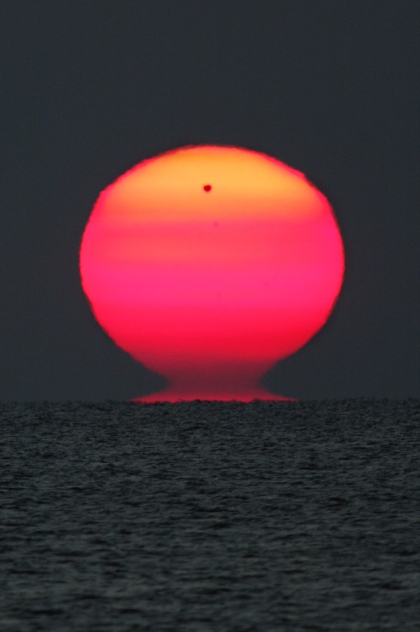 Die Sonne geht über dem Schwarzen Meer am Horizont auf. Oben ist ein schwarzer Punkt, es ist der Planet Venus. Die Sonne ist unten dunkelrot und oben hellorange, sie hat die Form einer etruskischen Vase.