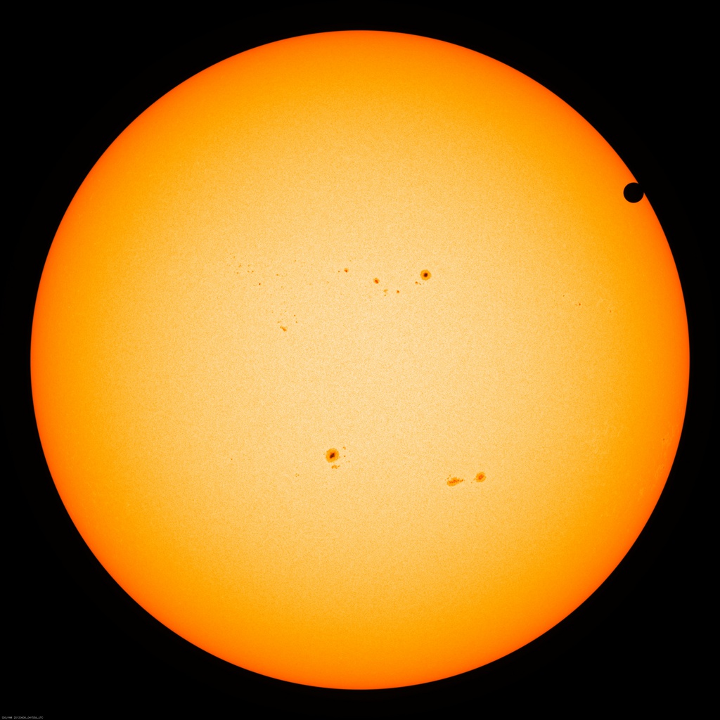 Die ganze Sonne füllt das Bild, in der Mitte leuchtet sie dunkelgelb, am Rand ist sie orangefarben. Auf der Oberfläche sind mehrere Sonnenflecken verteilt. Rechts oben ist eine schwarze runde Einkerbung, es ist der Planet Venus am Sonnenrand.