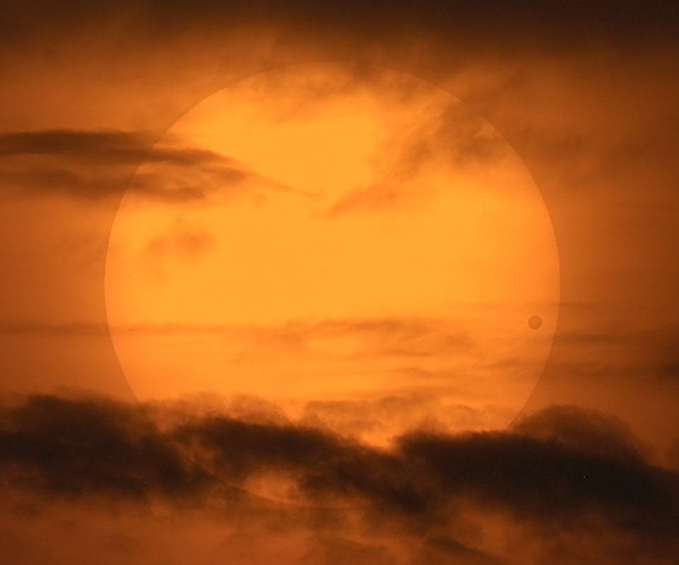 Hinter Dunst und Wolken leuchtet die orangefarbene Sonne. Rechts zeichnet sich ein dunkler Fleck ab, es ist der Planet Venus vor der Sonnenscheibe.