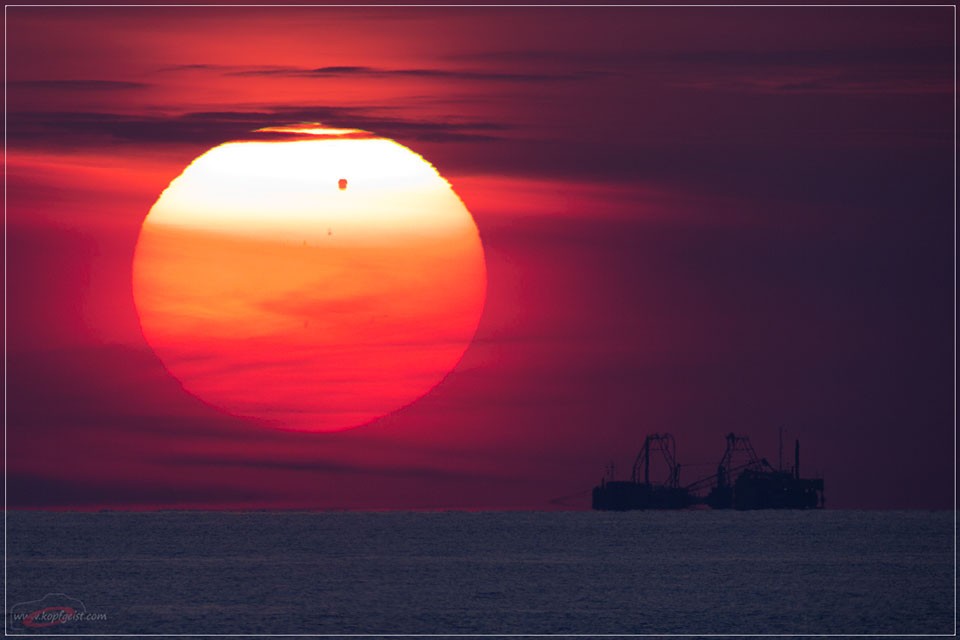 Über dem Meer leuchtet die orangerote Sonne hinter schleierartigen Wolken über dem Meer. Oben ist die Sonne noch weiß, dort ist der Fleck des Planeten Venus zu sehen. Recht unten am Horizont des Meeres sind die Silhouetten von Schiffen zu sehen.
