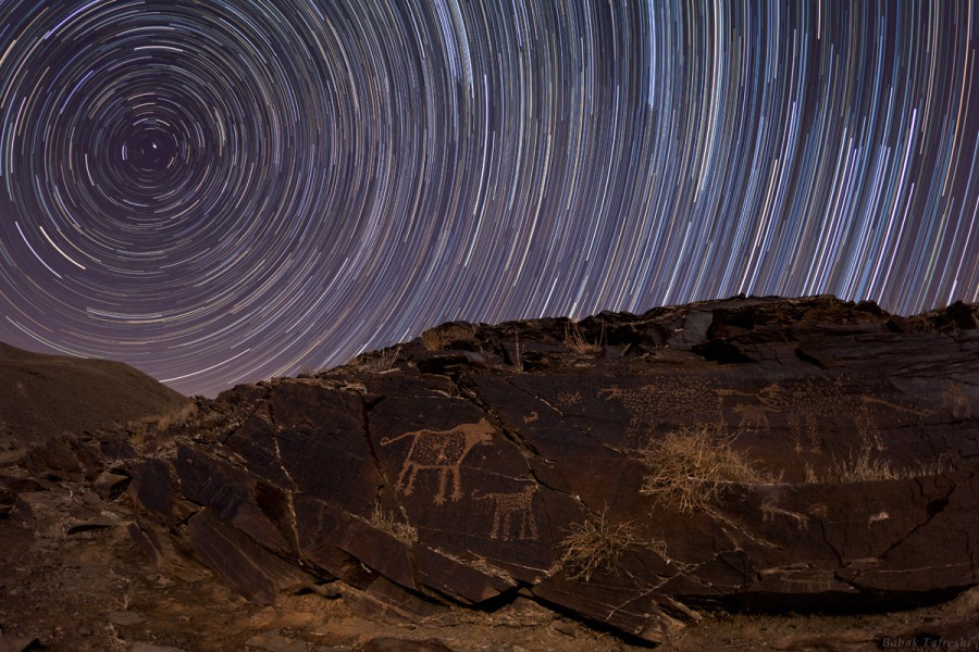 Hinter einer Felswand mit Petroglyphen sind Strichspuren am Himmel, links kreisen die Bögen um den Himmelsnordpol.