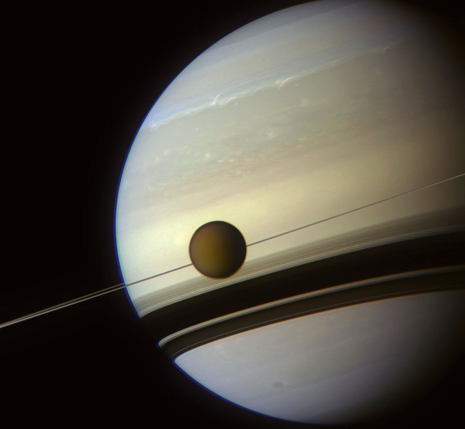 Von rechts ragt Saturn riesig ins Bild, das rechts an seinem Terminator endet. Die Wirbelstürme und Wolkenbänder sind in zarten blauen und gelben Farbtönen hervorgehoben. Die Ringe verlaufen schräg als dünne Linie durchs Bild, die Schatten fallen breit nach unten auf den Planeten. Mitten über den Ringen ist der wolkenbedeckte Mond Titan.