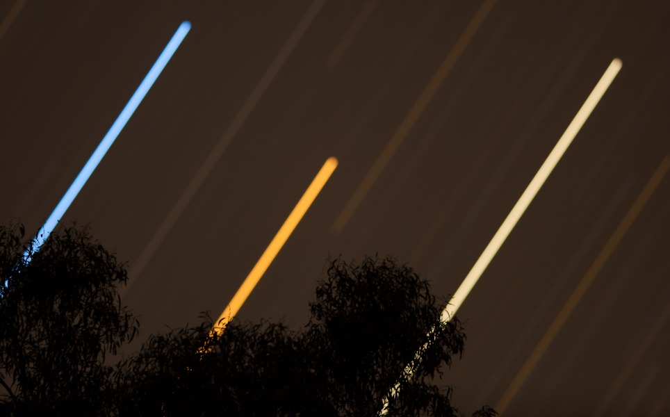 Die hellen Himmelslichter Spica, Mars und Saturn gehen zusammen hinter Bäumen unter. Ihre unscharfen Bilder ziehen farbige Spuren am Himmel.