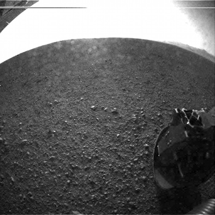 Das Fischaugen-Weitwinkelbild zeigt eine steinige Marslandschaft, in der rechten unteren Ecke ragt ein Rad des Rovers Curiosity mit einem Durchmesser von 50 Zentimetern ins Bild.