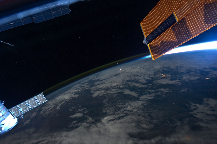 Der Blick fällt von der Internationalen Raumstation ISS auf die Erde. Rechts ragt ein Solarpaneel ins Bild, dahinter ist eine schmale Spitze der Erde beleuchtet, rechts unten ist eine Raumkapsel. Hinten krümmt sich der Erdrand mit grünlichem Nachthimmellicht, und in der Bildmitte blitzt ein Meteor auf.
