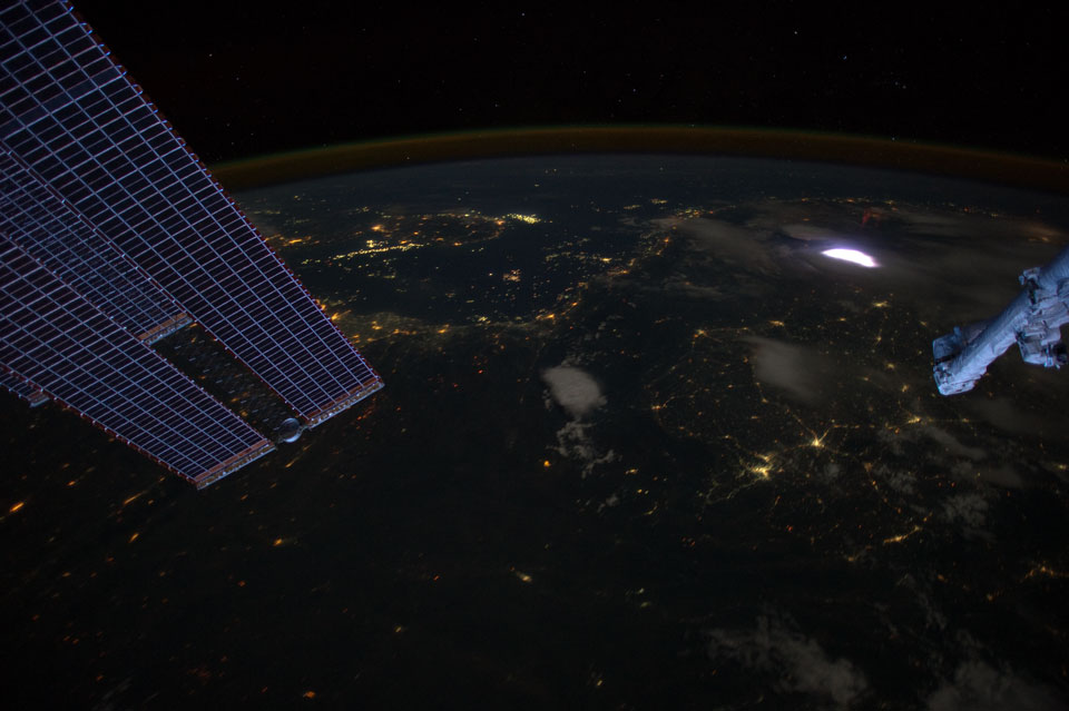 Hinter Solarpaneelen und dem Roboterarm der ISS ist die dunkle Erde mit beleuchteten Städten Wolken, Nachthimmellicht am Horizont, einem Gewitter und - kaum erkennbar - einem roten Kobold.