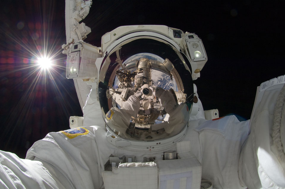 Ein Astronaut blickt in die Kamera, die er selbst hält. Hinter ihm leuchtet die Sonne. Im Visier spiegeln sich die Kamera, der Roboterarm, die ISS und die Erde.