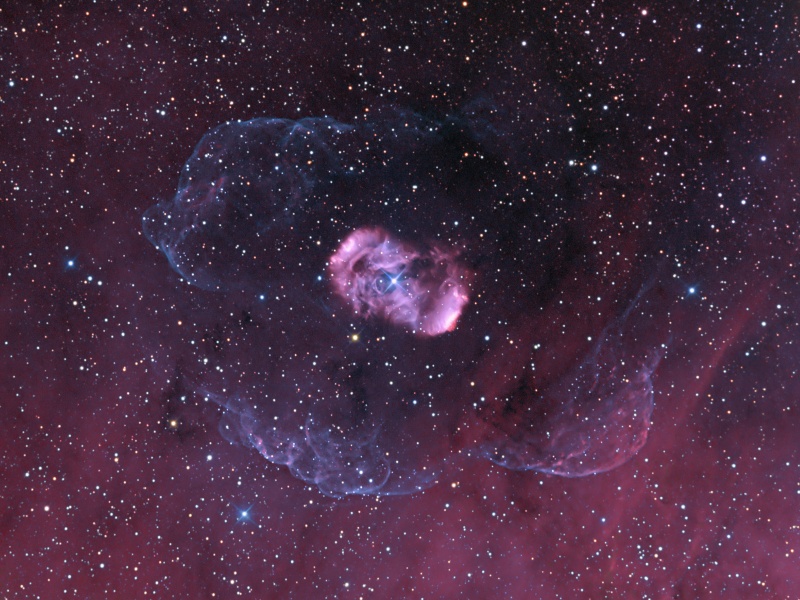 Mitten im Bild leuchtet vor einem matten purpurfarbenen Hintergrund ein kompakter zyklamefarbener Nebel, der von zarten blauen Schlieren umgeben ist.