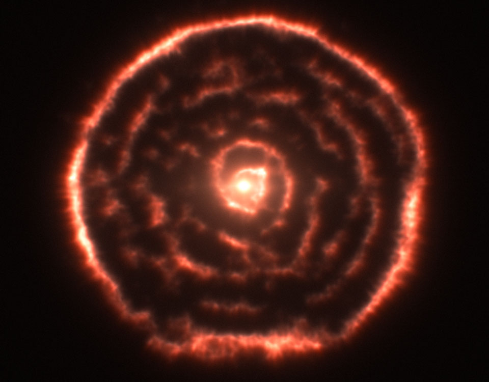 Um einen hellen Stern verläuft eine brüchige, leicht unregelmäßige Spirale in roten Farbtönen. Außen herum verläuft ein relativ heller Kranz.
