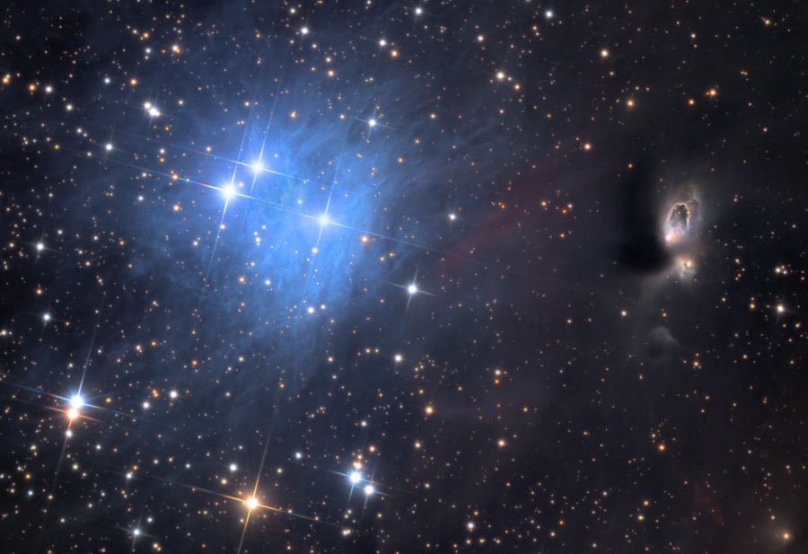 Im Bild mit vielen gezackten Sternen leuchtet links ein blauer Nebel, entfernt erinnert die Anordnung an die Plejaden.