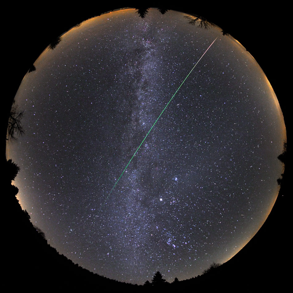 Das Bild zeigt den ganzen Himmel, am Rand verläuft der Horizont als Kreis. Quer durchs ganze Bild zieht ein Meteor eine grünliche Leuchtspur. Die Milchstraße verläuft senkrecht durchs Bild.