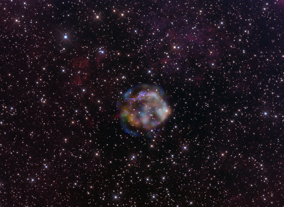 Mitten im Bild ist eine verschwommene, farbige Wolke, umgeben von einem gleichmäßigen Sternenfeld. Die Wolke ist der Überrest der Supernova Cas A.