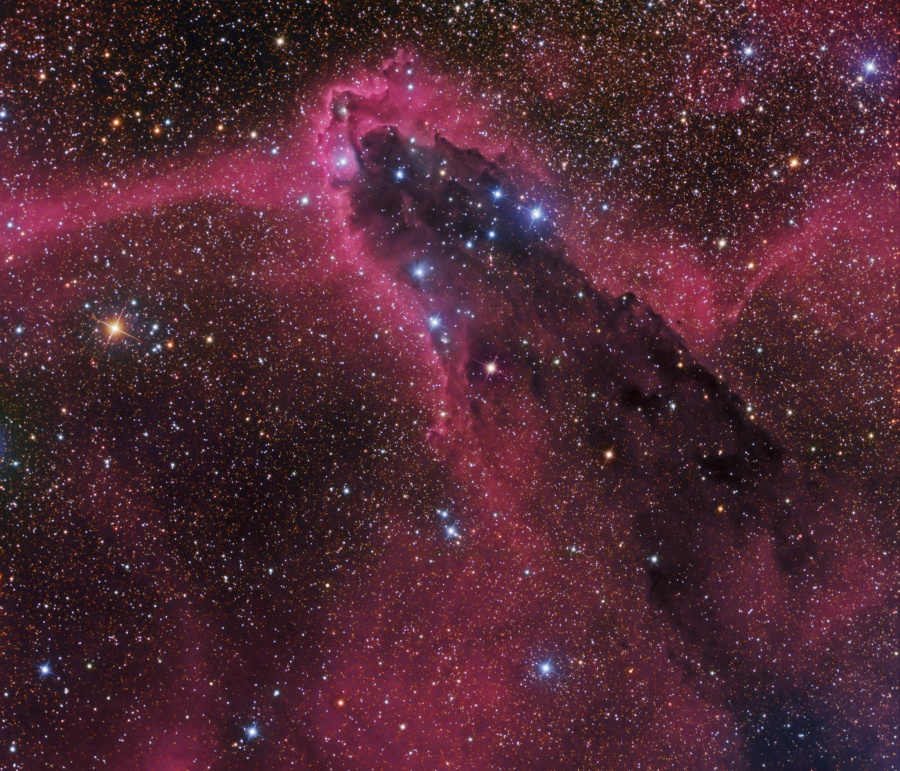 Von links unten ragt eine dunkle Struktur ins Bild, die von rot leuchtenden Nebeln umgeben ist. Am oberen Ende des dunklen Turms leuchtet eine Sterngruppe.