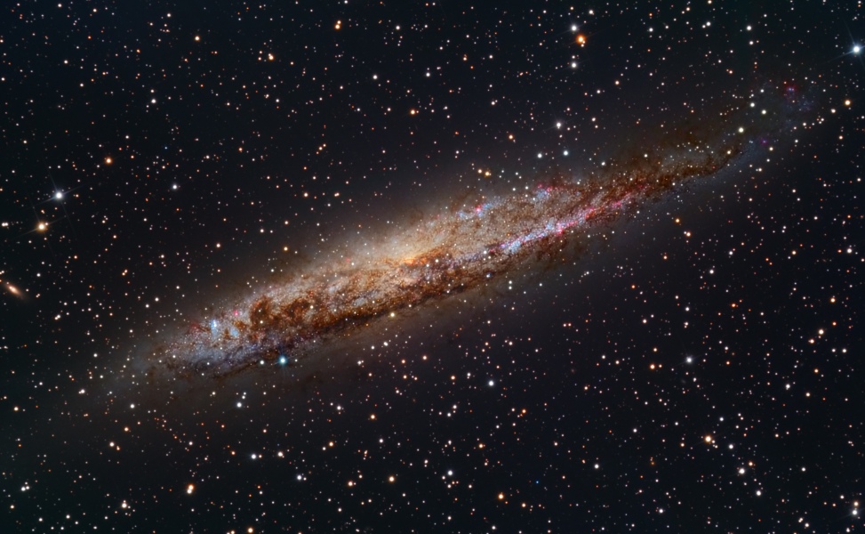 Schräg im Bild ist eine von vielen Sternen umgebene Spiralgalaxie. Sie wirkt etwas unruhig und asymmetrisch. Links leuchtet sie etwas heller. Sie ist von vielen dunklen Staubwolken überzogen. Sie ist von schräg oben fast von der Seite zu sehen.