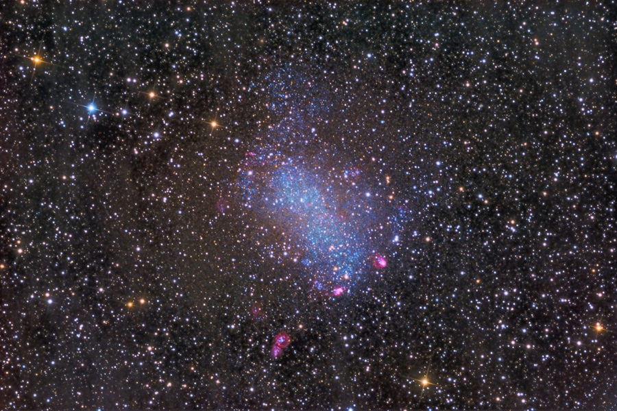 Mitten in einem dichten Sternfeld leuchtet eine Ansammlung von blauen Sternen und rosaroten Sternbildungsregionen. Sie gehören zu Barnards Galaxie, auch 