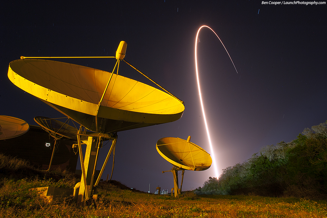 Hinter gelb beleuchteten Antennenschüsseln, die wie Pflanzen aus dem Boden wachsen, steigt der Bogen einer startenden Atlas-V-Rakete zum Himmel auf.