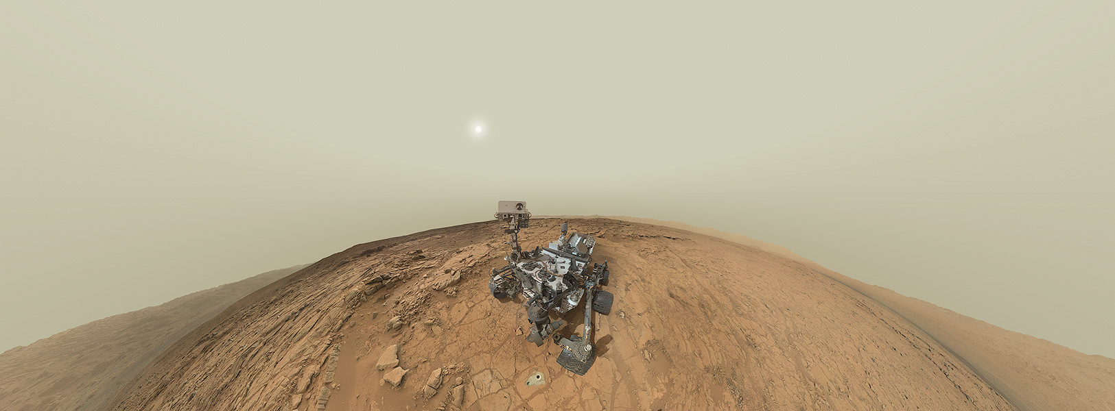 Mitten in einer stark gekrümmten Landschaft hockt ein Marsrover. Der Arm, mit dem die Einzelbilder aufgenommen wurden, ist nicht abgebildet. Im Hintergrund leuchtet die blasse Sonne hinter Nebelschleiern.