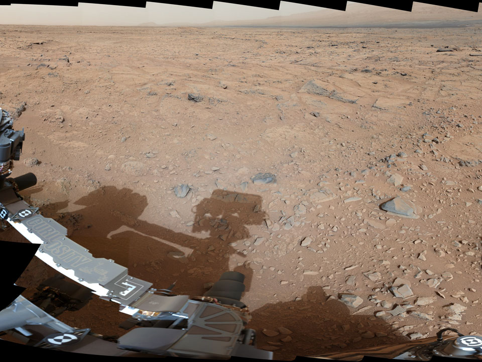 Das Panorama zeigt die Weite der Marslandschaft, vorne links ragt der Rover Curiosity ins Bild und wirft einen Schatten.