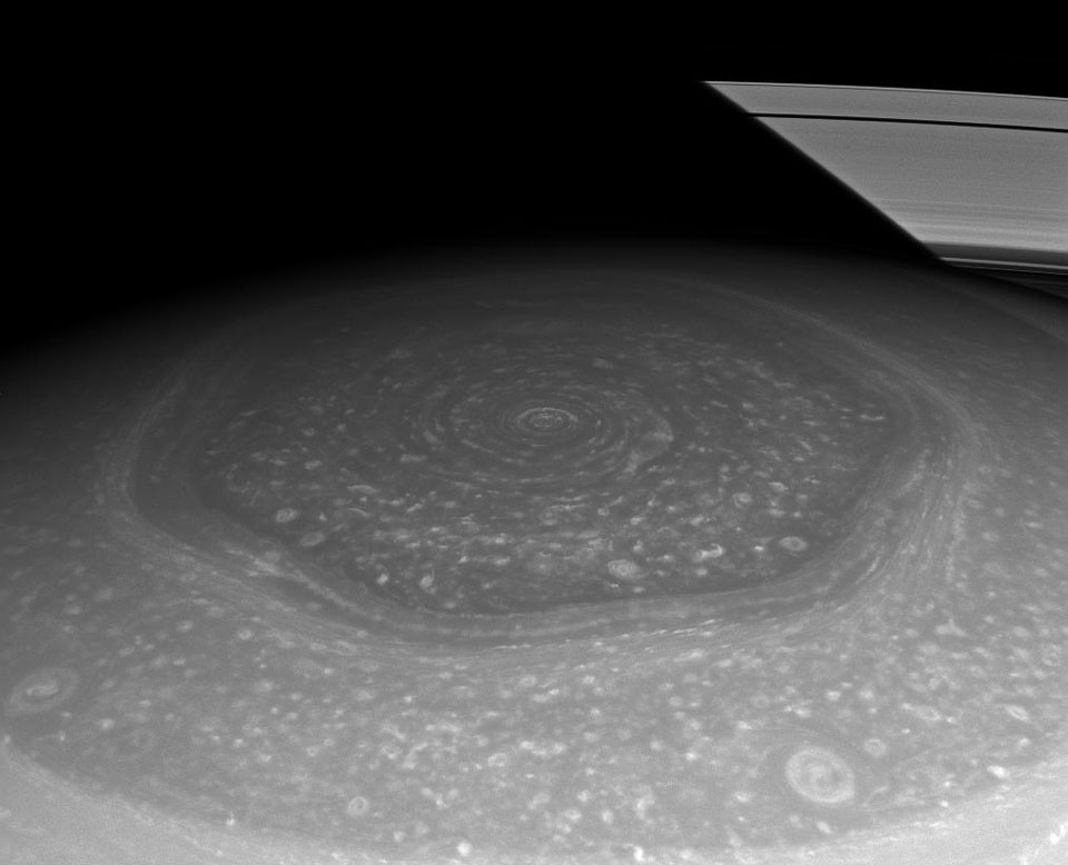 Mitten im Bild ist eine seltsame Wolkenstruktur um Saturns Nordpol abgebildet. Sie sieht wie ein regelmäßiges Sechseck aus. Hinten ist ein kleiner Teil der Ringe, auf die der Schatten des Planeten fällt.