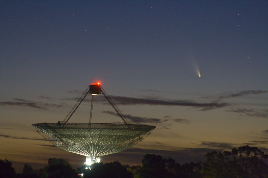 Links zeigt die Schüssel einer Radioantenne zum Himmel, oben leuchtet ein rotes Licht beim Empfänger. Rechts über dem Teleskop steht Komet PanSTARRS (C/2011 L4), sein Schweif zeigt nach oben von der Sonne weg. Die Sonne ist bereits untergegangen.