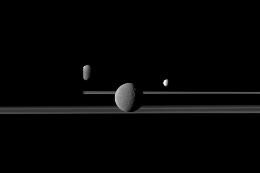 Drei Monde schweben über den Ringen des Saturn. Links verschwindet ein Mond sowie der hintere Teil der Ringe hinter dem dunklen Saturn, der nicht im Bild erkennbar ist.