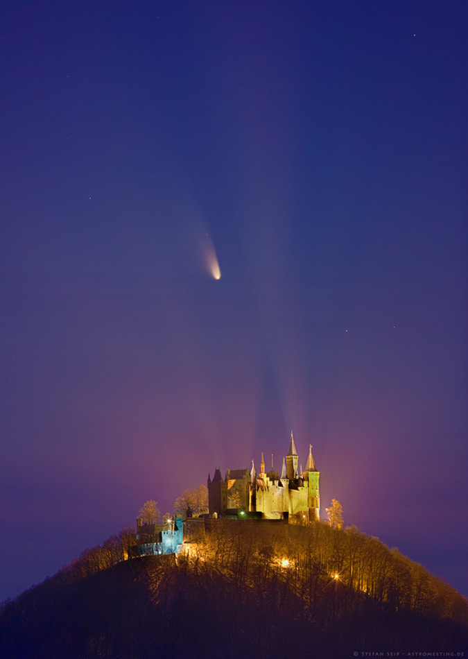 Über einem hell beleuchteten malerischen Kometenschloss auf der Kuppe eines blattlosen Waldes leuchtet ein Komet. Die Scheinwerfer, die das Schloss beleuchten, werfen Lichtkegel an den Himmel.