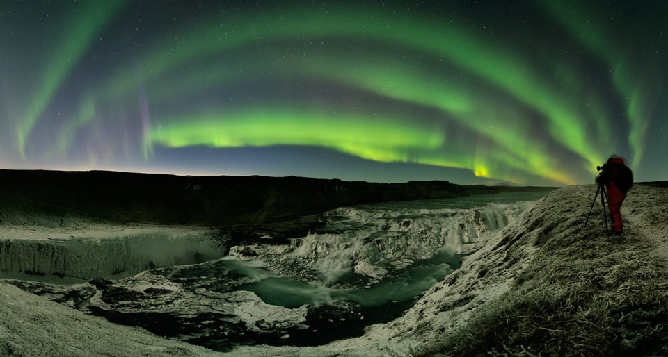 Über einem isländischen Wasserfall wölbt sich ein geschichtetes grünes Polarlicht. Links ist der Komet PanSTARRS eine leichte Einkerbung am Horizont.