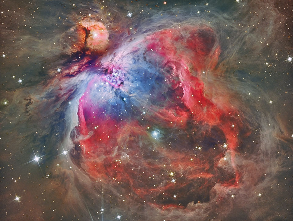 Der Orionnebel zeichnet sich bunt in einer dunkelgrauen Nebeltapete ab. Die Höhlung mit dem Trapez links oben ist blau gefärbt, die Ranken darunter leuchten tiefrot.